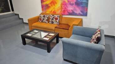Uma sala de estar moderna apresenta um sofá laranja, poltrona azul, almofadas florais, mesa de centro de vidro com decoração e uma pintura abstrata vibrante na parede.