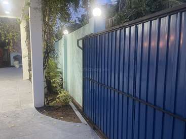 Um portão de metal azul fica ao lado de uma parede azul clara com lâmpadas esféricas brilhantes. Nas proximidades, vegetação e videiras sobem, formando uma cena serena de entrada residencial.