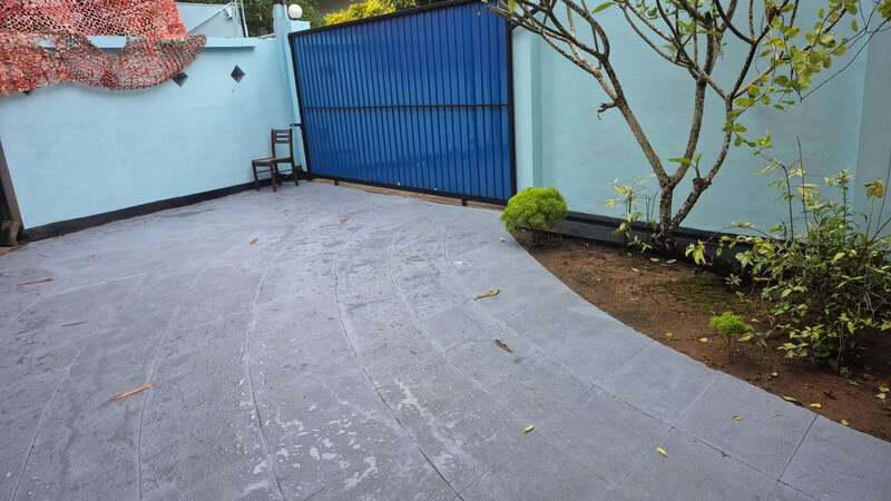 Esta imagem mostra um pátio limpo e pavimentado com paredes azuis, um portão azul, uma cadeira, uma árvore, pequenas plantas e algumas folhas caídas no chão.