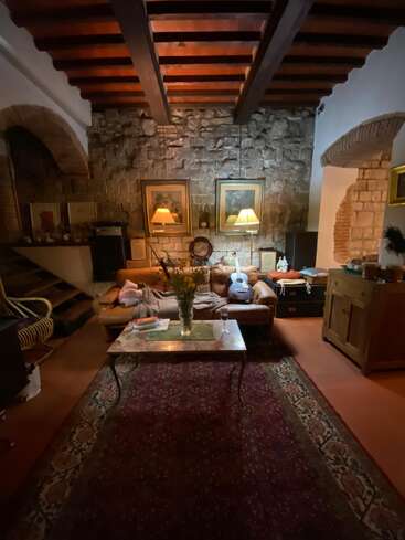 Un salon chaleureux avec des murs en pierre, des poutres apparentes, un éclairage tamisé, un canapé en cuir, une guitare, des fleurs sur la table basse, des livres et un grand tapis à motifs. Une ambiance accueillante.