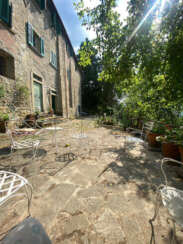 La lumière du soleil inonde une terrasse rustique en pierre, meublée de chaises en métal blanc et entourée d'une végétation luxuriante. Un vieux bâtiment en pierre aux volets verts ajoute au charme des lieux. Une atmosphère paisible.