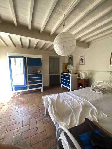 Cette chambre chaleureuse est dotée de poutres apparentes, d'un sol en terre cuite, d'un lit en métal blanc aux draps froissés, d'armoires bleues et blanches, de meubles en bois et d'un éclairage assuré par des lanternes en papier.