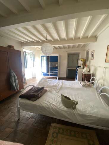 Une chambre chaleureuse avec un plafond à poutres apparentes blanches, un lit double, une armoire en bois, une commode bleue, des fenêtres ouvertes, de la lumière naturelle, un sol rustique et une décoration sobre.