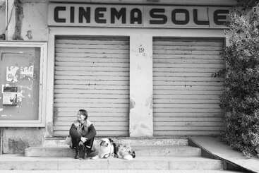 Une femme est assise sur les marches avec son chien, devant un cinéma fermé et aux volets baissés, appelé « Cinema Sole ». La scène est en noir et blanc.
