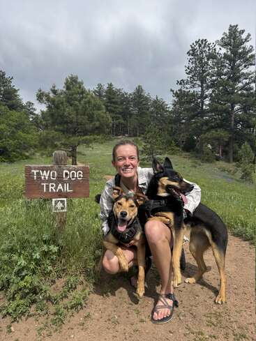 Uma mulher sorridente está ajoelhada ao lado de dois cachorros felizes, perto de uma placa de madeira com os dizeres “Two Dog Trail”, cercada por uma vegetação exuberante e pinheiros, sob um céu nublado.