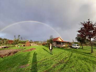 Une personne se tient sur de l'herbe verte sous un large arc-en-ciel, près d'une petite maison au toit rouge, d'arbres, d'une camionnette et d'un ciel nuageux dramatique.