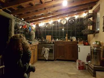 Une cuisine rustique et confortable avec des armoires en bois, des casseroles suspendues et une personne assise au premier plan. Un éclairage chaud met en valeur les ustensiles de cuisine bien organisés et les éléments essentiels de la cuisine.