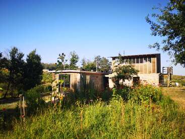 Une maison moderne et écologique se dresse au milieu d'une végétation luxuriante, de fleurs sauvages et d'herbes hautes. Entourée d'arbres, elle se prélasse sous un ciel bleu clair, rayonnant de tranquillité.