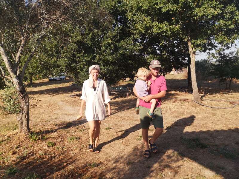 Une femme et un homme portant un enfant marchent dehors sur un sol sec. Les arbres donnent de l'ombre et l'atmosphère est lumineuse, chaude et détendue.