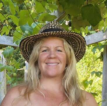 Eine Frau mit langen blonden Haaren lächelt freundlich, trägt einen Strohhut und steht an einem sonnigen Tag draußen, umgeben von grünen Pflanzen und Holzgittern.
