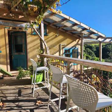 The image depicts a serene wooden deck with white chairs, a bar, and a house in the background, set against a clear blue sky and lush greenery.