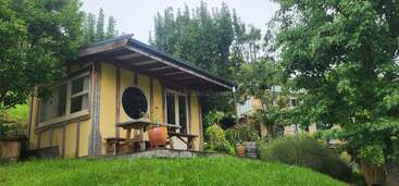 A cozy, small yellow house with a circular window sits on a grassy hill. Surrounded by trees and plants, it has a wooden porch and benches.