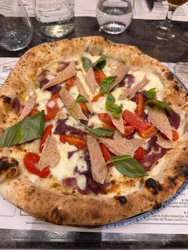 Uma pizza recém-assada está sobre a mesa, coberta com queijo derretido, tomates cereja, folhas de manjericão, fatias de carne e rodeada por copos e cardápios.