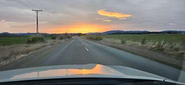 A imagem mostra uma estrada rural com uma borda gramada, com um carro se afastando da câmera, tendo como pano de fundo árvores e um céu claro.