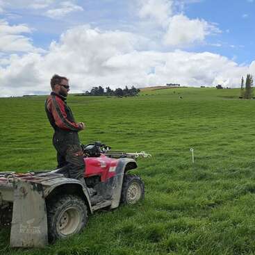 La imagen muestra a un hombre de pie sobre un vehículo todoterreno en un campo cubierto de hierba, con una alambrada y vacas lejanas visibles bajo un cielo nublado.