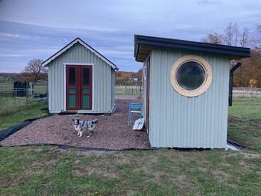 Two small pastel green sheds stand on gravel, one with a round window. A blue chair and an Australian Shepherd dog appear in the foreground. Rural landscape.