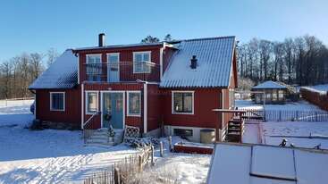 A red wooden house with white trim stands in a snowy landscape. There is a balcony, steps to the door, and bare trees surrounding the property.