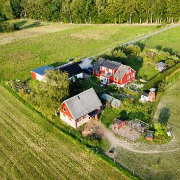 This aerial image shows a charming rural farmstead with red houses, greenhouses, animal enclosures, gardens, and surrounding fields bordered by trees and a winding road.