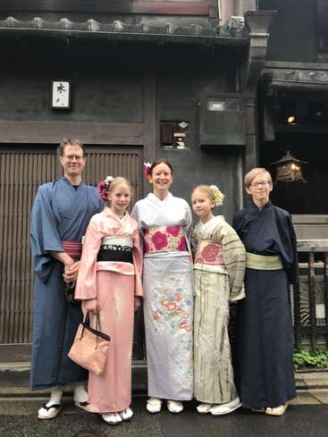 A family of five poses in front of a traditional Japanese building, all wearing beautiful kimonos with floral patterns, geta sandals, and hair accessories.
