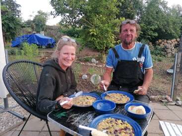 Um homem e uma mulher sorridentes estão sentados ao ar livre, à mesa, saboreando uma refeição juntos. Eles têm tigelas azuis com comida, rodeados por um jardim verdejante.