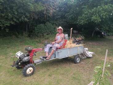 Um homem sorridente, usando um chapéu de cowboy, está sentado em um trator artesanal com um reboque cheio de palha, cercado por vegetação em um cenário rural ao ar livre.