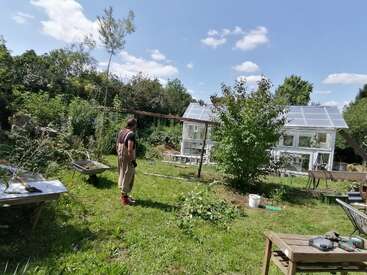Uma pessoa está em um jardim verdejante e exuberante, perto de uma estufa, num dia ensolarado. Ferramentas, um carrinho de mão e uma mesa estão espalhados por ali.