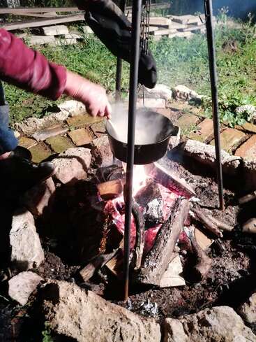 Uma pessoa está mexendo uma panela suspensa sobre uma fogueira, cercada por pedras e tijolos, com luvas penduradas acima. A cena se passa ao ar livre, à noite.