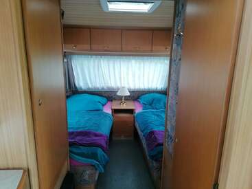 Esta imagem mostra um quarto aconchegante de trailer com duas camas de solteiro, roupa de cama em tons de azul-petróleo e roxo, uma mesinha de cabeceira com um abajur, armários de madeira e uma janela com cortina.