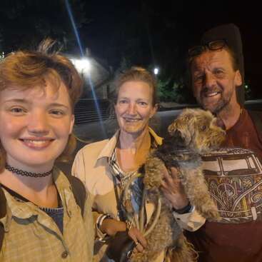 Tres personas posan felices juntas por la noche al aire libre; una de ellas sostiene en brazos a un perrito pequeño y peludo. Todos sonríen con calidez, disfrutando del momento bajo la luz de las farolas.