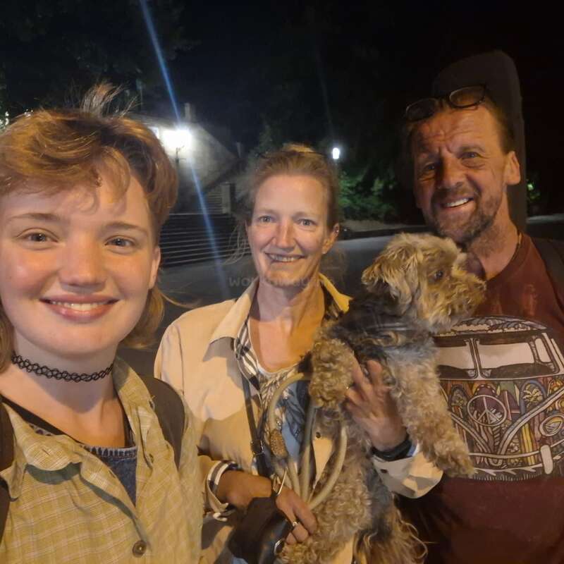 Três pessoas posam juntas, felizes, à noite ao ar livre, com uma delas segurando um cachorrinho fofinho. Todas sorriem calorosamente, aproveitando o momento sob a luz dos postes.