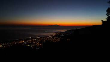 This image captures a stunning coastal city at dusk, with city lights glowing under a vibrant orange and blue sunset sky, and mountains silhouetted in the background.