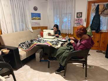 Two women sit in a cozy room, working on textiles—one uses a sewing machine, the other hand-sews. Fabrics and sewing supplies cover the table.
