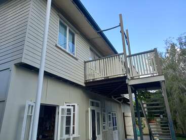 The image depicts the rear of a two-story house featuring a balcony, staircase, and a garage, with trees visible in the background on a sunny day.