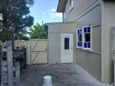 The image depicts the side of a house with a white door and window, a fence, and a concrete patio, surrounded by trees and a blue sky.