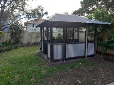 The image depicts a small, open-sided wooden structure with a corrugated metal roof and walls, situated in a backyard surrounded by trees and a fence.