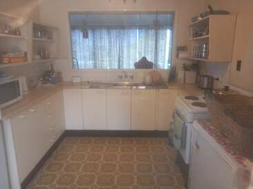 The image depicts a compact kitchen with white cabinets and countertops, featuring a sink, stove, and refrigerator, with a window above the sink offering a view of the outside.