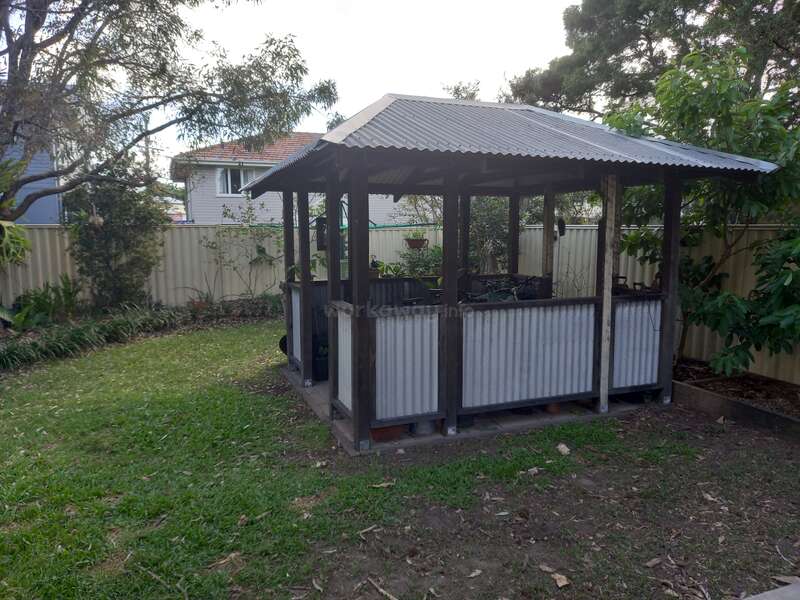 The image depicts a small, open-sided wooden structure with a corrugated metal roof and walls, situated in a backyard surrounded by trees and a fence.