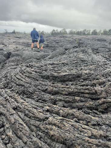 Deux personnes vêtues de vestes bleues traversent un champ de lave volcanique sombre et accidenté sous un ciel nuageux, avec des arbres visibles à l'horizon.