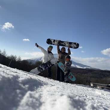 The image depicts two individuals standing on a snowy hill, each holding a snowboard, with one person extending their arm in a celebratory gesture.