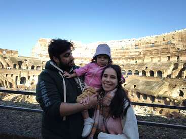 Uma família de três pessoas posa dentro do Coliseu, em Roma, sorrindo sob um céu azul claro. O pai segura o filho pequeno, e todos aproveitam o passeio turístico.