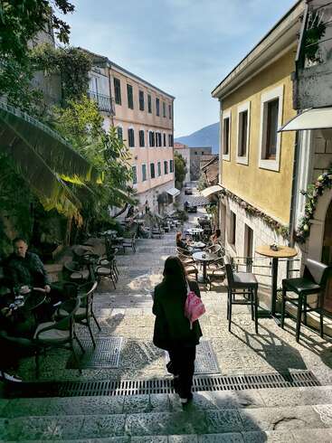 Uma mulher com uma mochila rosa caminha por uma rua pitoresca de pedra, cheia de cafés ao ar livre, prédios coloridos, vegetação e montanhas distantes sob um céu claro.