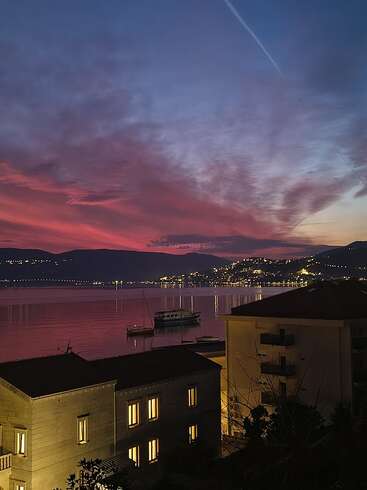 Uma cidade costeira deslumbrante ao entardecer, com nuvens roxas e alaranjadas vibrantes, águas calmas e reflexivas, barcos, janelas iluminadas e luzes cintilantes nas colinas distantes.