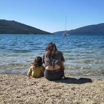 A woman and child sit on a pebbled beach, gazing at the calm sea. A sailboat floats in the distance, surrounded by green hills. Peaceful scene.
