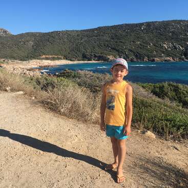 Ein kleiner Junge in einem gelben Tanktop, blauen Shorts und einer Mütze steht auf einem Feldweg, hinter ihm erstrecken sich eine malerische Küste und Hügel.
