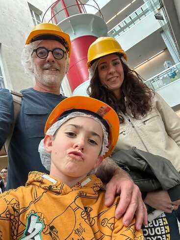 Eine dreiköpfige Familie posiert drinnen für ein Selfie, alle tragen gelbe Schutzhelme und Haarnetze und stehen vor einer großen rot-weißen Konstruktion.