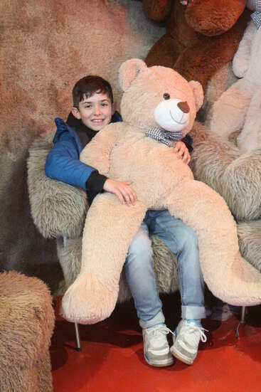 Ein lächelnder Junge in einer blauen Jacke sitzt auf einem Plüschsessel, drückt einen großen Teddybären an sich, ist von anderen Plüschbären umgeben und sieht fröhlich aus.