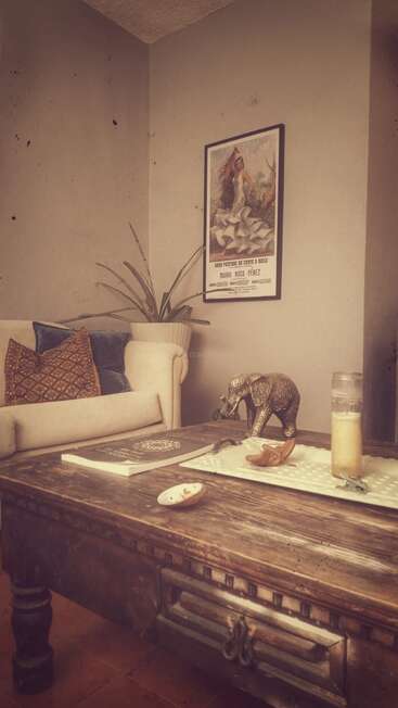 Uma sala de estar aconchegante com um sofá creme, almofadas coloridas, planta de interior, mesa de madeira vintage, estatueta de elefante, vela e um pôster de dança espanhola emoldurado na parede.