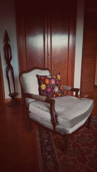 Uma poltrona vintage com estofamento claro e estrutura de madeira fica sobre um tapete estampado, com uma almofada floral colorida, perto de portas de madeira e uma escultura abstrata.