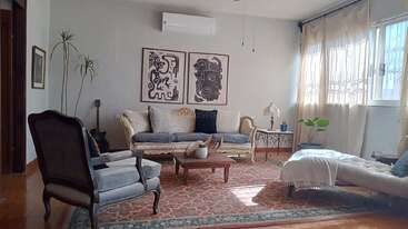 Esta sala de estar apresenta um sofá vintage, poltronas, tapete estampado, obras de arte modernas, plantas, janelas grandes com cortinas, uma mesa de centro e uma decoração aconchegante e convidativa.