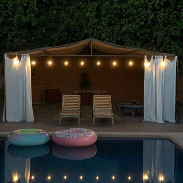 L'image représente une arrière-cour sereine avec une piscine, deux chaises longues et un gazebo orné de guirlandes lumineuses, entouré d'arbres luxuriants et d'un ciel bleu clair.